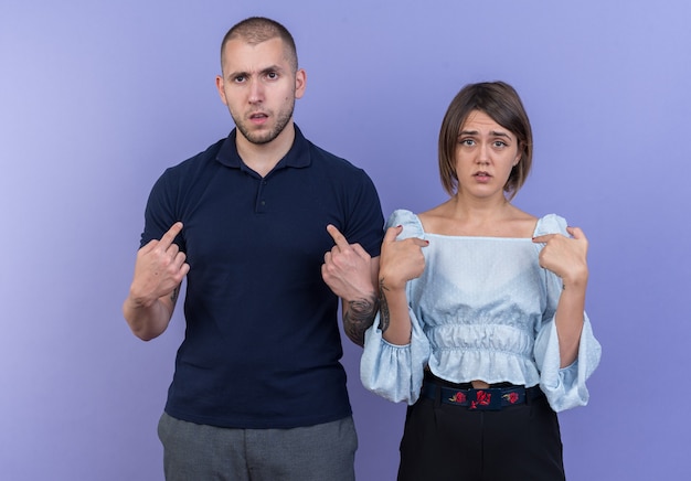 Hermosa joven pareja hombre y mujer confundidos apuntando a sí mismos de pie sobre la pared azul