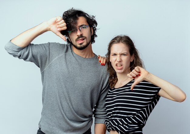 Hermosa joven pareja hombre y mujer disgustados mostrando los pulgares hacia abajo sobre la pared blanca