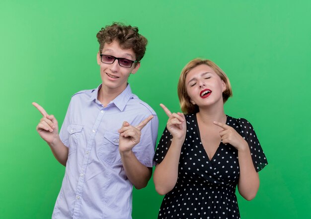 Hermosa joven pareja hombre y mujer feliz y positivo apuntando con los dedos hacia el lado de pie sobre la pared verde