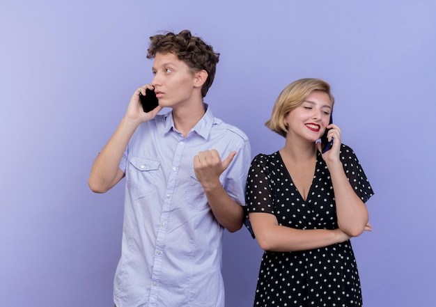 Hermosa joven pareja hombre y mujer hablando por teléfono móvil de pie sobre la pared azul