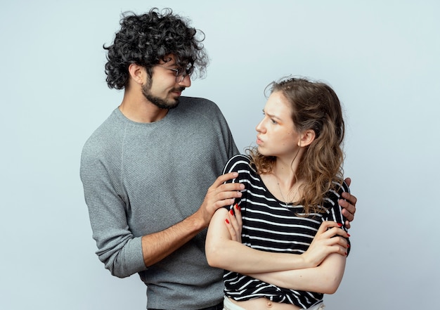 Hermosa joven pareja hombre y mujer, hombre pidiendo perdón mujer disgustada después de la pelea de pie con los brazos cruzados sobre la pared blanca