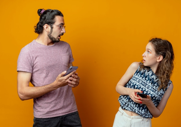 Hermosa joven pareja hombre y mujer mirando el uno al otro con expresión confusa mientras sostiene teléfonos inteligentes de pie sobre la pared naranja