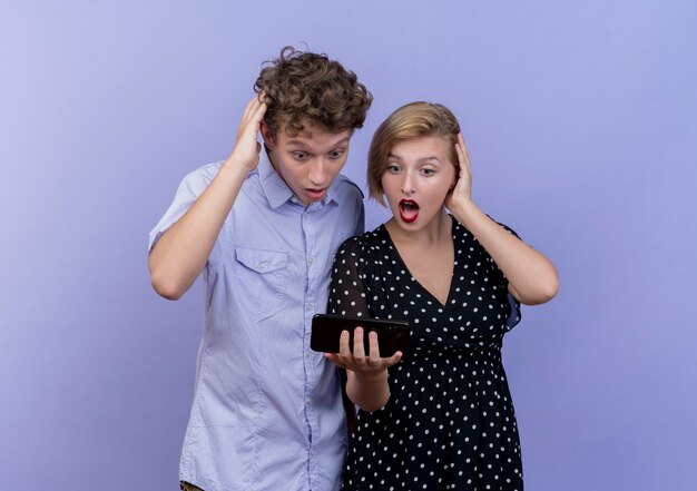 Hermosa joven pareja hombre y mujer mirando la pantalla del teléfono inteligente sorprendido y asombrado de pie sobre la pared azul