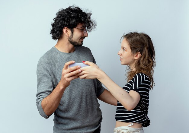 Foto gratuita hermosa joven pareja hombre y mujer mirándose sosteniendo un teléfono inteligente tomando una foto de ellos juntos de pie sobre la pared blanca