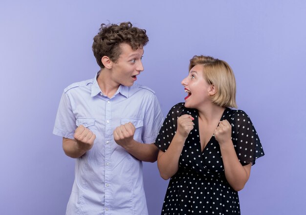 Hermosa joven pareja hombre y mujer de pie uno al lado del otro apretando los puños felices y emocionados sobre la pared azul