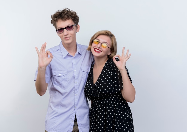 Foto gratuita hermosa joven pareja hombre y mujer sonriendo mostrando signo ok de pie sobre la pared blanca