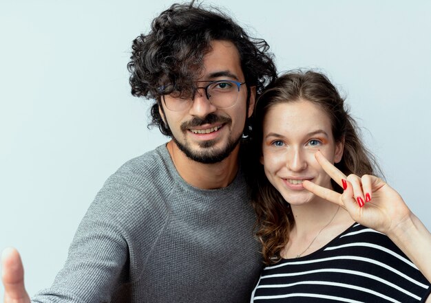 Hermosa joven pareja hombre y mujer sonriendo, mujer mostrando el signo de la victoria sobre la pared blanca