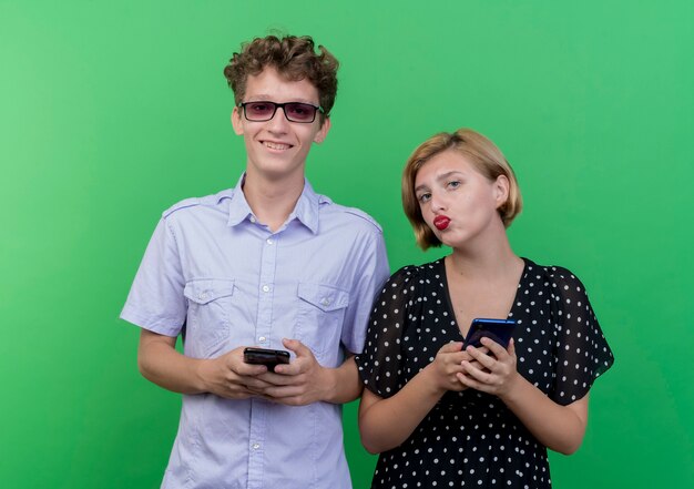 Hermosa joven pareja hombre y mujer sosteniendo smartphones sonriendo de pie sobre la pared verde