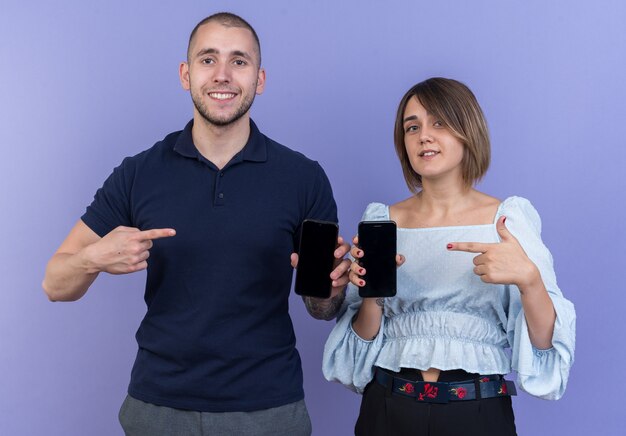 Hermosa joven pareja hombre y mujer sosteniendo teléfonos inteligentes apuntando con el dedo índice a los teléfonos sonriendo alegremente feliz y positivo de pie