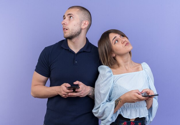 Hermosa joven pareja hombre y mujer con teléfonos inteligentes mirando hacia arriba con miradas de ensueño pensando en posición positiva sobre la pared azul