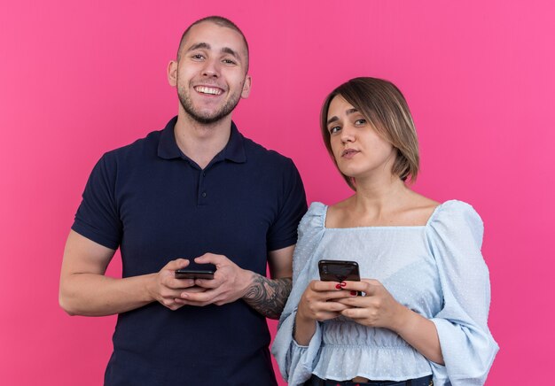 Hermosa joven pareja hombre y mujer con teléfonos inteligentes sonriendo alegremente de pie sobre la pared rosa