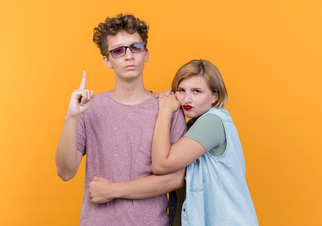 Hermosa joven pareja hombre y mujer vistiendo ropa casual hombre mostrando el dedo índice mientras su novia disgustaba de pie sobre la pared naranja