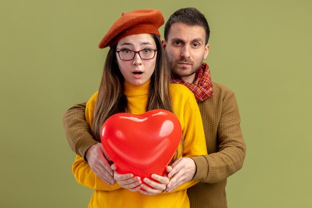 Hermosa joven pareja mujer en boina con globo en forma de corazón y hombre con bufanda alrededor del cuello sorprendido celebrando el día de San Valentín de pie sobre la pared verde