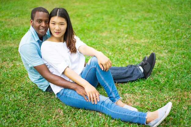 Hermosa joven pareja multiétnica abrazando y sentado en la hierba verde.