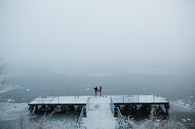 Hermosa joven pareja de pie en un muelle y mira el lago congelado