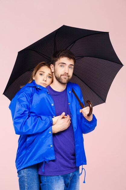 Foto gratuita hermosa joven pareja posando vistiendo una capa de lluvia con paraguas sobre pared rosa claro