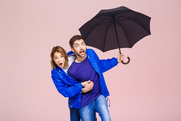 Hermosa joven pareja posando vistiendo una capa de lluvia con paraguas sobre pared rosa claro