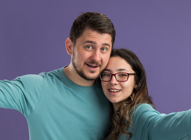 Hermosa joven pareja en ropa casual azul hombre y mujer sonriendo alegremente felices enamorados juntos de pie sobre la pared púrpura