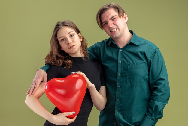 Hermosa joven pareja en ropa casual confundido hombre y mujer positiva con globo en forma de corazón celebrando el día de San Valentín de pie sobre la pared verde