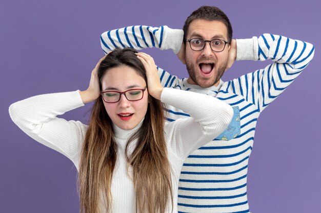 Hermosa joven pareja en ropa casual feliz y sorprendido hombre y mujer tomados de la mano en la cabeza concepto de día de San Valentín de pie sobre la pared púrpura