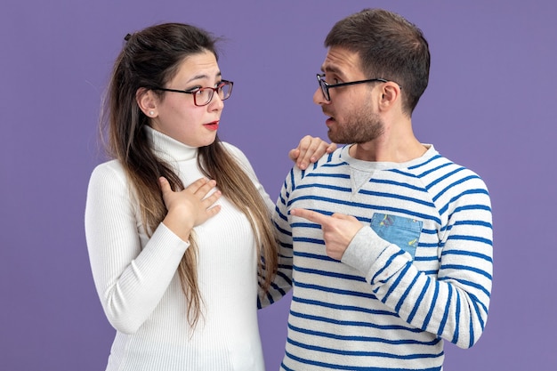 Hermosa joven pareja en ropa casual hombre confundido apuntando con el dedo índice a su novia sorprendida celebrando el día de San Valentín de pie sobre la pared púrpura