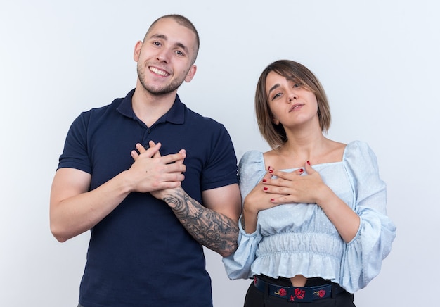 Hermosa joven pareja en ropa casual hombre y mujer feliz y positivo tomados de la mano en el pecho sintiéndose agradecido de pie sobre la pared blanca