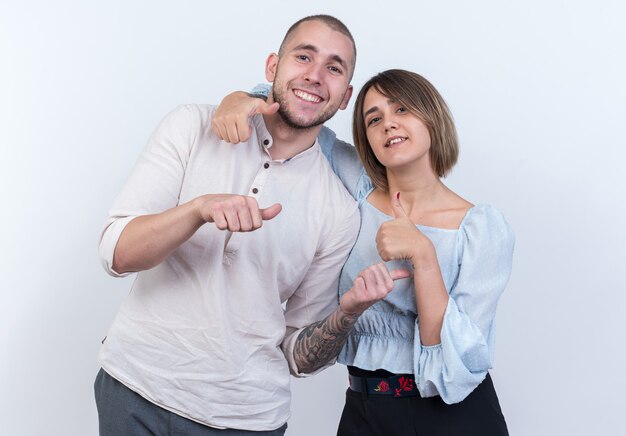 Hermosa joven pareja en ropa casual hombre y mujer sonriendo alegremente mostrando los pulgares para arriba feliz y positivo de pie sobre la pared blanca