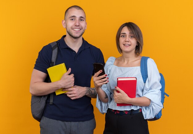 Hermosa joven pareja en ropa casual con mochilas sosteniendo libros y smartphone mirando sonriendo alegremente feliz y positivo de pie