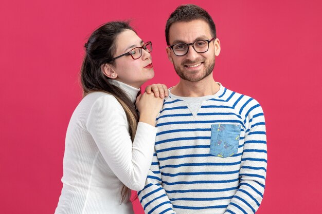 Hermosa joven pareja en ropa casual mujer feliz va a besar a su novio sonriente celebrando el día de San Valentín de pie sobre la pared rosa