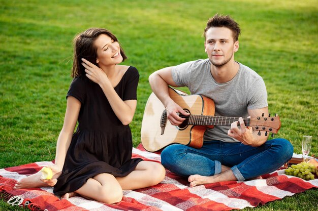 Hermosa joven pareja sonriendo, descansando, relajándose en un picnic en el parque. Hombre tocando la guitarra.