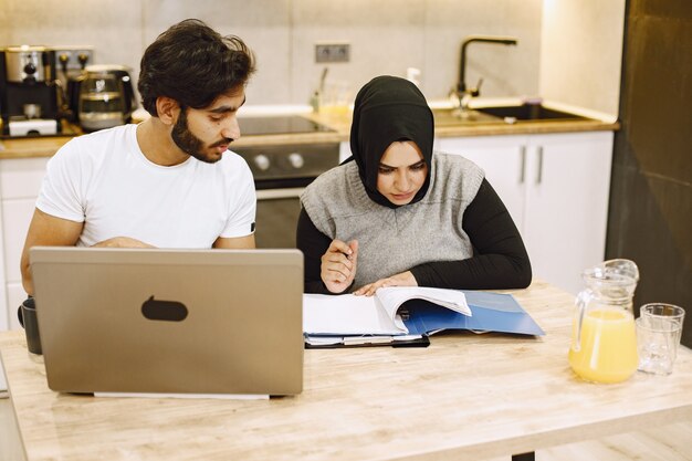 Hermosa joven pareja usando una computadora portátil, escribiendo en un cuaderno, sentado en una cocina en casa. Chica árabe con hidjab negro.