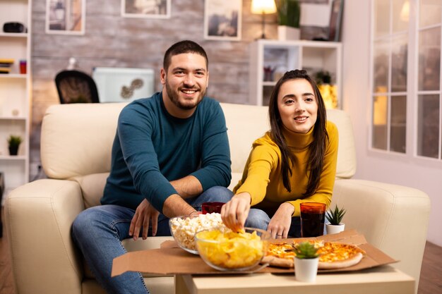 Hermosa joven pareja viendo la televisión y comiendo comida rápida para llevar en la sala de estar