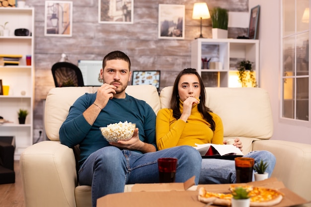 Hermosa joven pareja viendo la televisión y comiendo comida rápida para llevar en la sala de estar