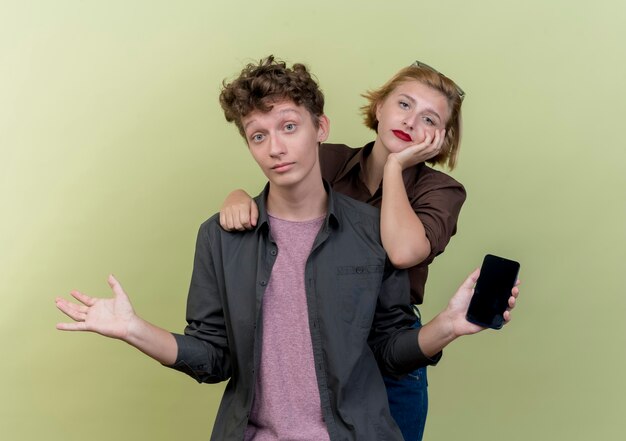 Hermosa joven pareja vistiendo ropa casual confundido hombre sosteniendo smartphone encogiéndose de hombros y su novia disgustada de pie sobre la pared de luz