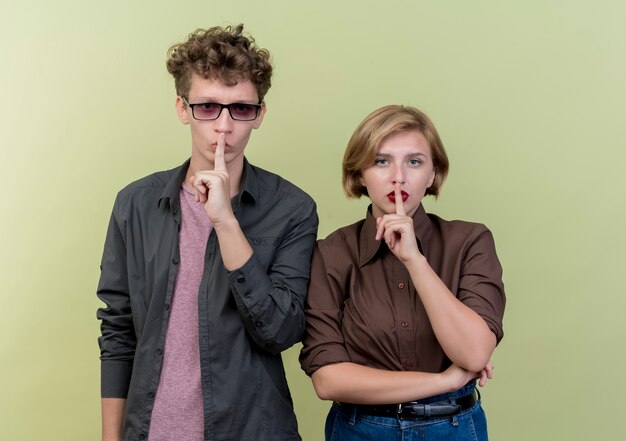 Hermosa joven pareja vistiendo ropa casual haciendo gesto de silencio con los dedos en los labios de pie sobre la pared de luz