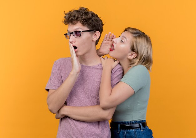 Hermosa joven pareja vistiendo ropa casual mujer susurrando un secreto a su novio sorprendido de pie sobre la pared naranja