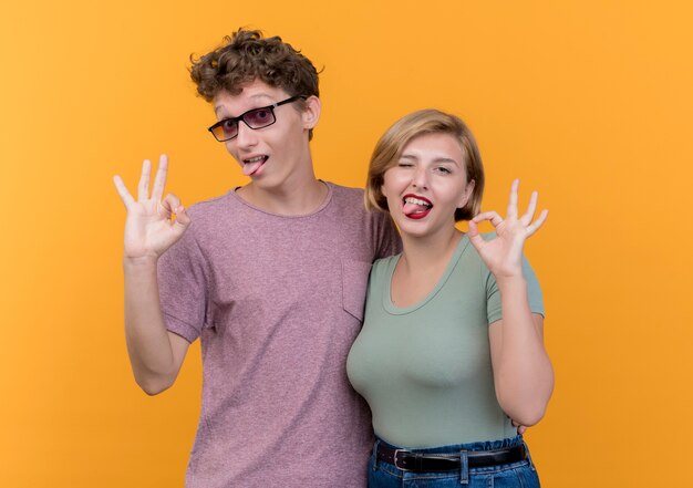 Hermosa joven pareja vistiendo ropa casual niño y niña posando y sonriendo mostrando signo ok de pie sobre la pared naranja