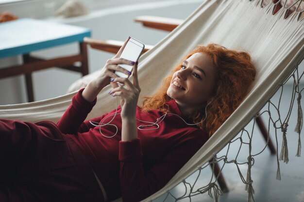 Hermosa joven pelirroja feliz se encuentra en una hamaca y chateando