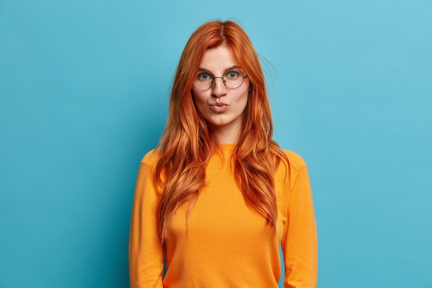 La hermosa joven pelirroja con gafas redondas mantiene los labios doblados quiere besar a alguien vestido con un jersey naranja que mira directamente.