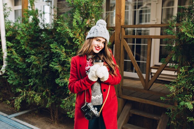 Hermosa joven con pelo largo en abrigo rojo y gorro de punto en casa de madera. Ella sostiene el café para llevar con guantes blancos, sonriendo amigablemente.