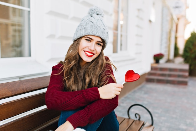 Hermosa joven con pelo largo en gorro de punto sentado en un banco en la calle. Ella tiene corazón de caramelo, sonriendo.