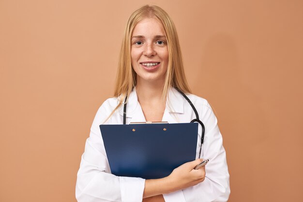 Hermosa joven practicante con experiencia en bata quirúrgica blanca con portapapeles