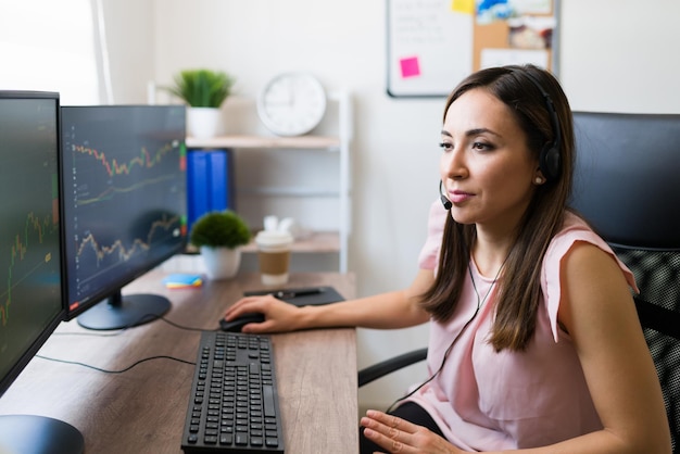 Hermosa joven que usa auriculares para llamar a los clientes mientras mira el monitor de la computadora y revisa el mercado de valores