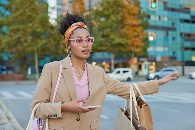 Hermosa joven rizada adicta a las compras atrapa el coche en la carretera sostiene el teléfono móvil en la mano lleva bolsas de papel hace compras en la ciudad