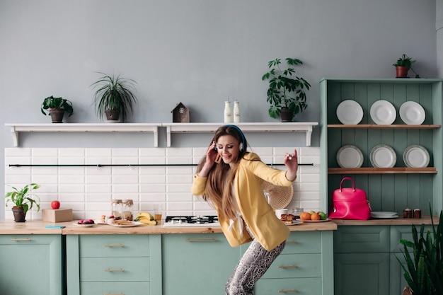 Hermosa joven en ropa de moda divirtiéndose en la cocina Modelo morena feliz con cabello largo escuchando música y bailando Mujer elegante con chaqueta amarilla y auriculares azules cantando