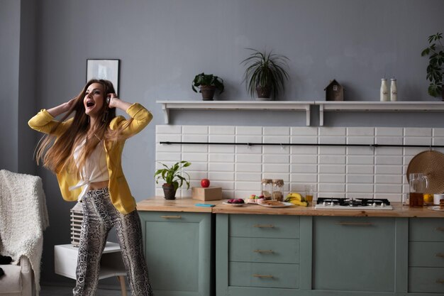 Hermosa joven en ropa de moda divirtiéndose en la cocina Modelo morena feliz con cabello largo escuchando música y bailando Mujer elegante con chaqueta amarilla y auriculares azules cantando