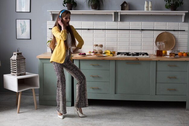 Hermosa joven en ropa de moda divirtiéndose en la cocina Modelo morena feliz con cabello largo escuchando música y bailando Mujer elegante con chaqueta amarilla y auriculares azules cantando