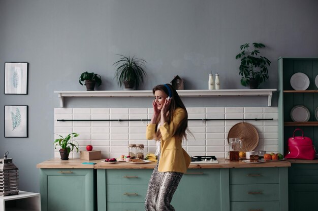 Hermosa joven en ropa de moda divirtiéndose en la cocina Modelo morena feliz con cabello largo escuchando música y bailando Mujer elegante con chaqueta amarilla y auriculares azules cantando