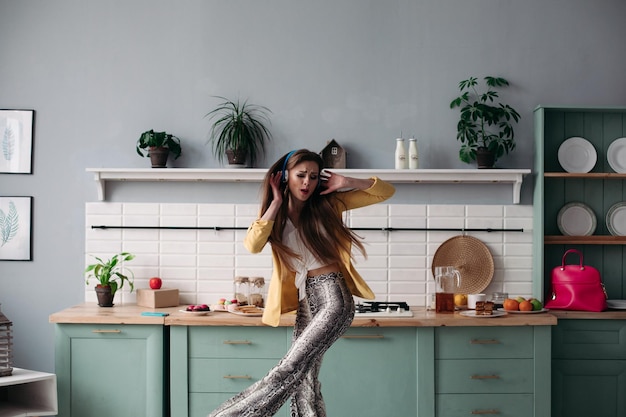 Foto gratuita hermosa joven en ropa de moda divirtiéndose en la cocina modelo morena feliz con cabello largo escuchando música y bailando mujer elegante con chaqueta amarilla y auriculares azules cantando