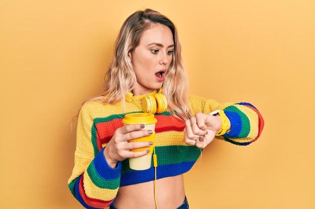 Hermosa joven rubia bebiendo una taza de café con auriculares mirando la hora del reloj preocupada, temerosa de llegar tarde